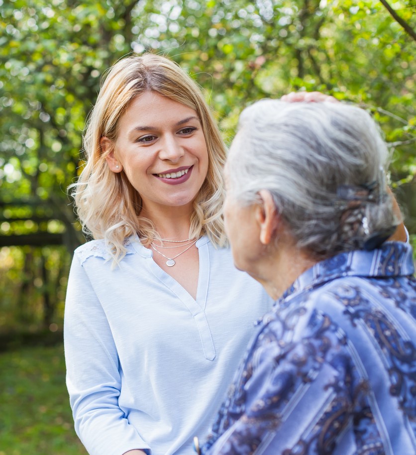 A young woman smiling and placing a hand on the shoulder of an older woman with gray hair in a sunlit outdoor setting.