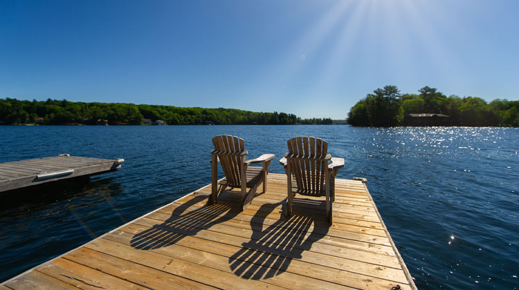 Cottage life - Sunrise on two empty Adirondack chairs sitting on a dock on a lake in Muskoka, Ontario Canada. The sun light create long shadows on the wooden pier.