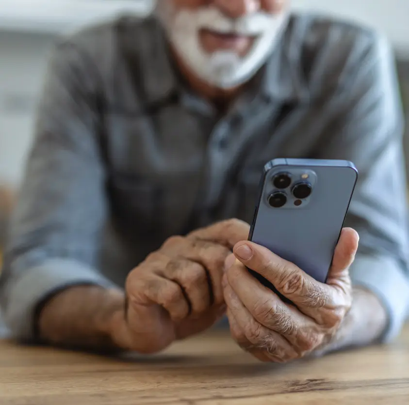 Close-up of an older man's hands holding a smartphone over a wooden table
